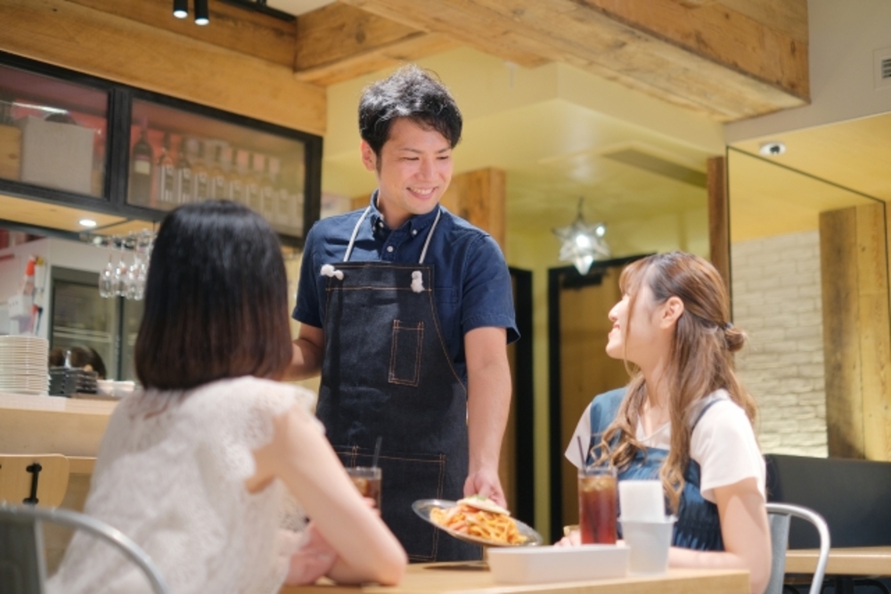 飲食店で女性2人に接客をする男性店員。料理を出す時に中国語で接客フレーズを添えると観光客から喜ばれる。